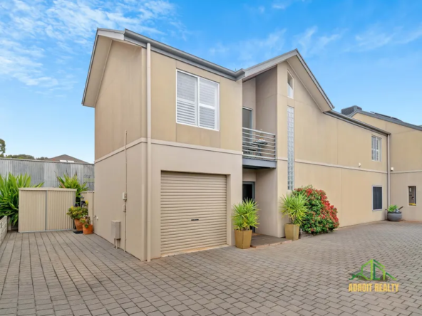Modern two-story tan townhouse with a single-car garage, upper-level balcony, and paved driveway featuring potted plants.