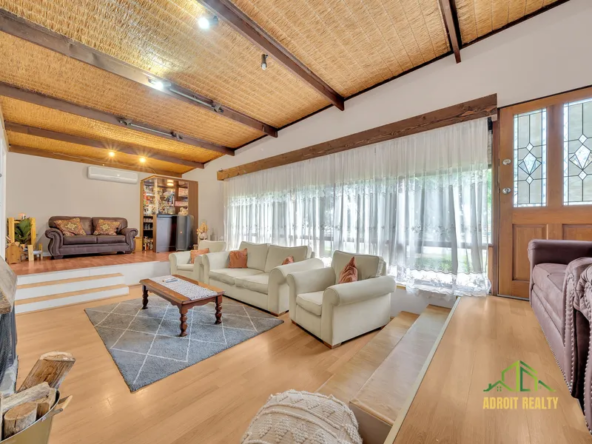 Mid-century style sunken living room featuring light wood flooring, exposed timber beams with a woven textured ceiling, and large windows with sheer white curtains.