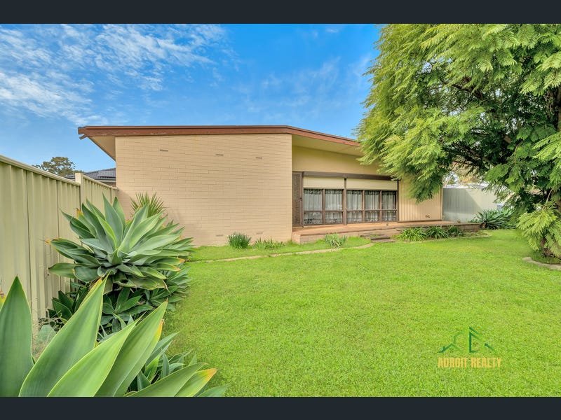 Exterior view of a tan brick home with a large green lawn, succulents, and a mature tree under a bright sky.